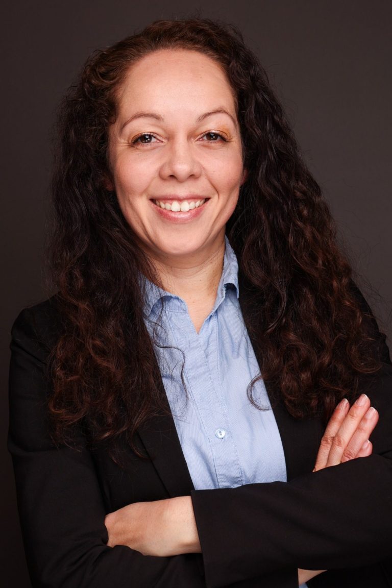 Lächelnde Frau mit lockigem Haar in beruflicher Kleidung, Hände verschränkt.